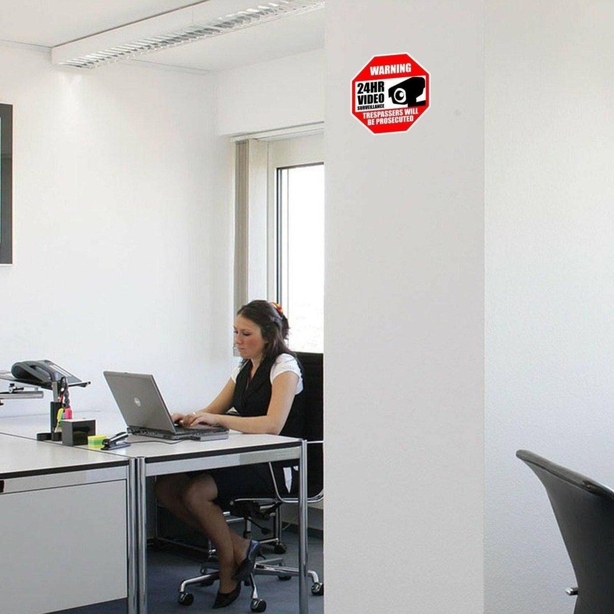 Office scene showing a woman working on a laptop with a video surveillance warning sign on the wall.