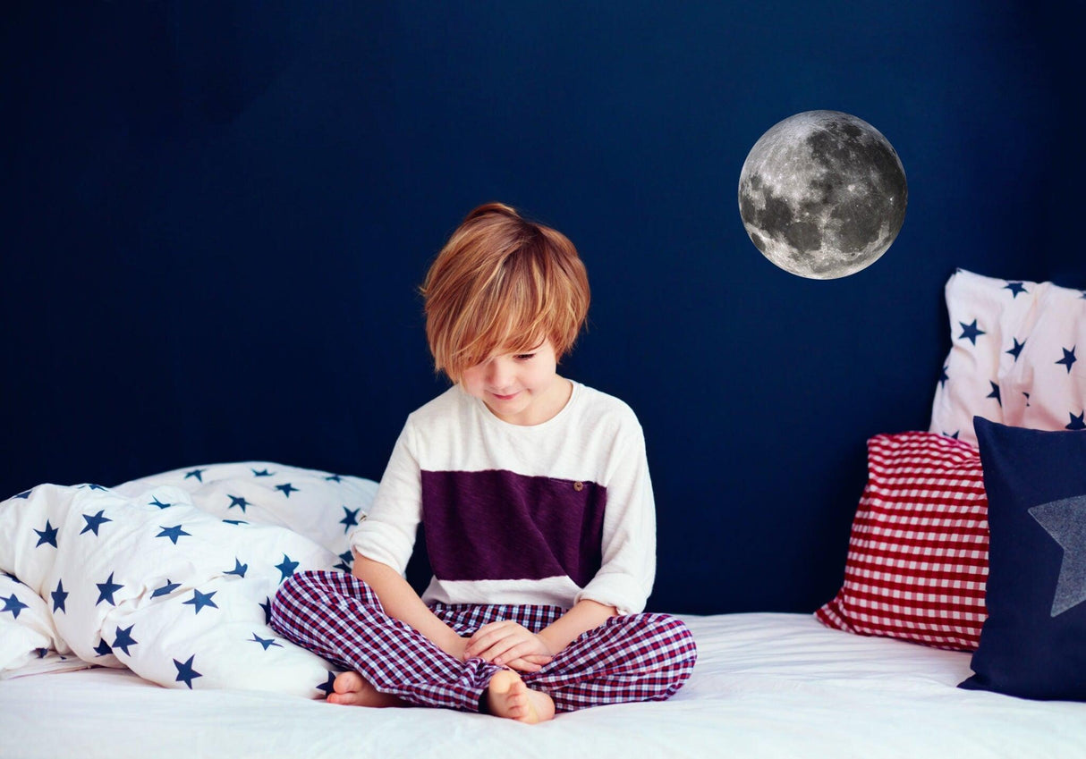 Boy sitting on bed decorated with star-themed bedding, with a full moon wall decal above.