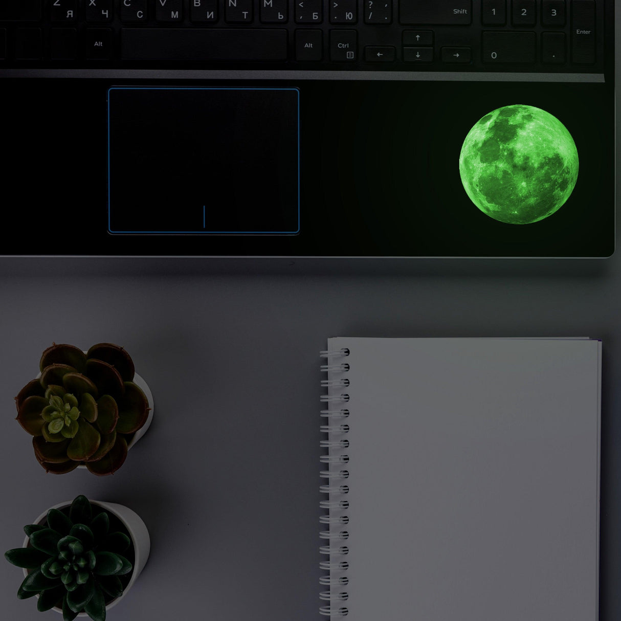 Lunar Luminescence laptop decal sticker glowing on laptop surface with succulents and notepad.