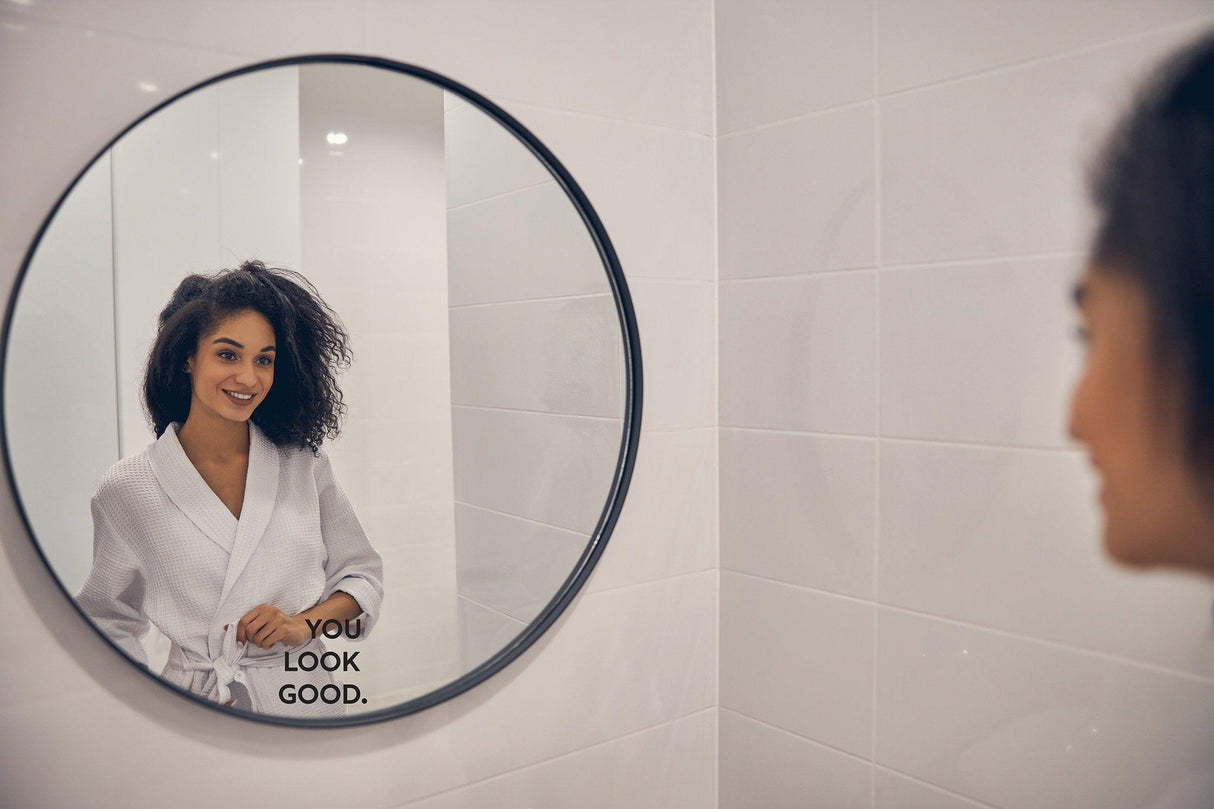 Woman smiling in a bathrobe, looking at a round mirror with 'You Look Good' decal.