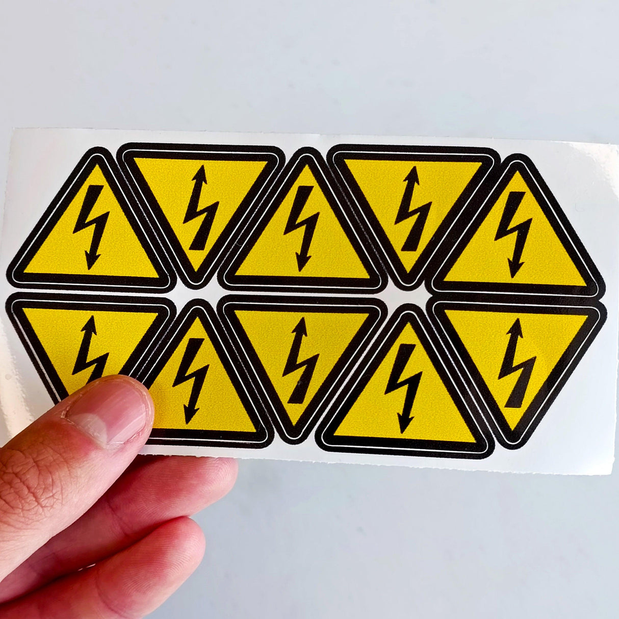 High Voltage Electrical Shock Hazard Stickers - Pack of 10 yellow triangle warning vinyl decals held in hand.