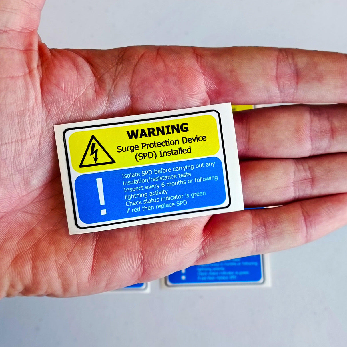Surge Protection Device warning label in hand, yellow and blue electrical safety instructions, SPD maintenance precautions.