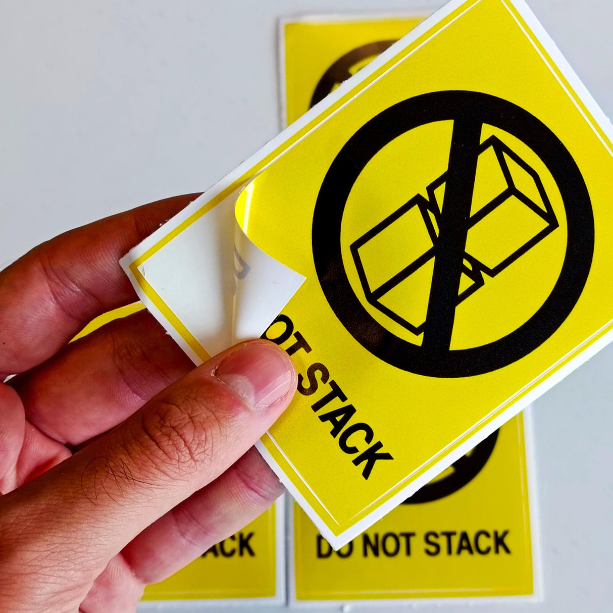 Hand holding a bright yellow Do Not Stack warning label sticker, designed for safe shipping and warehouse use.