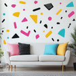 Colorful geometric Memphis pattern wall decals on a white wall above a modern couch with colorful pillows.