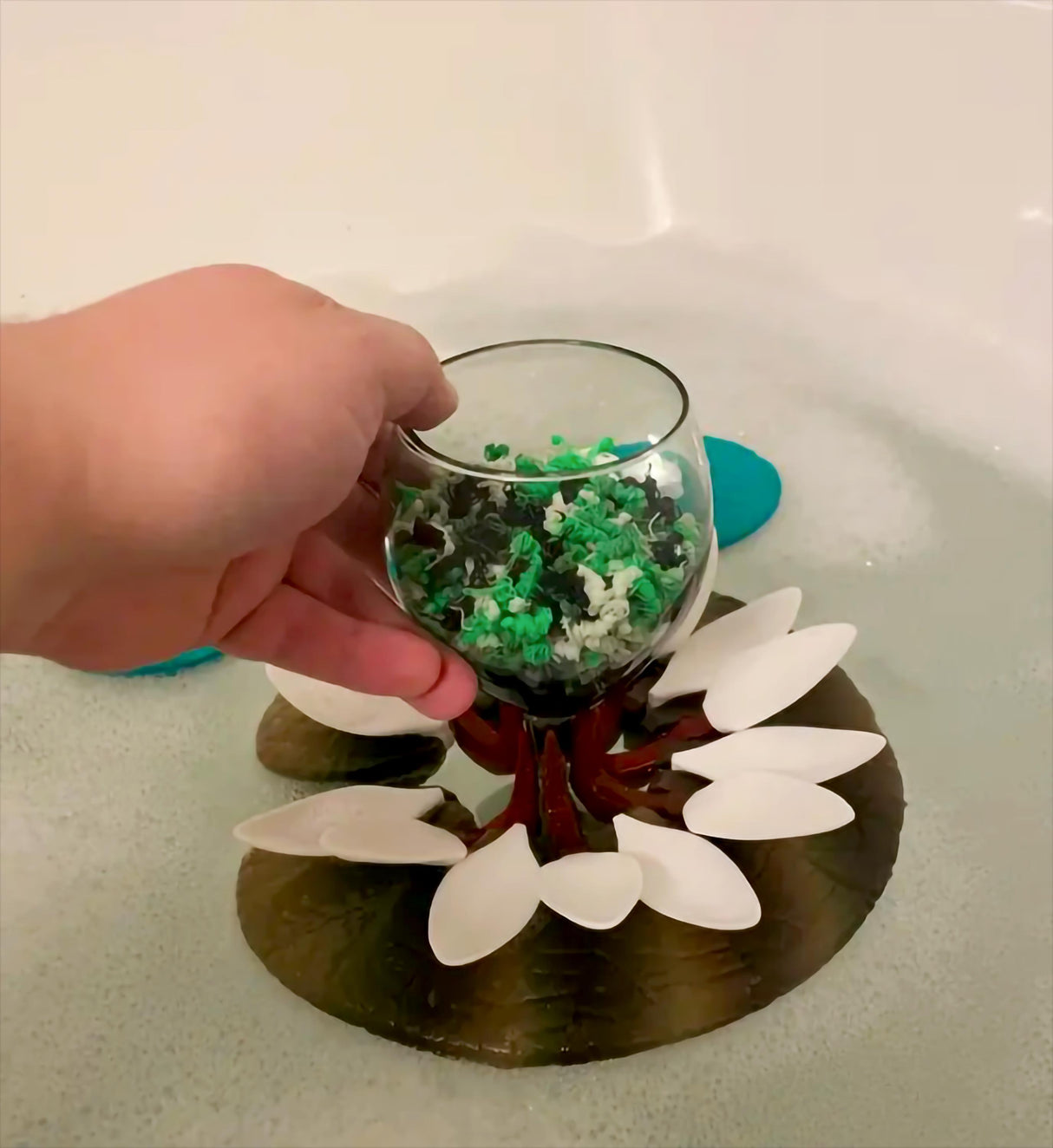 Hand holding a wine glass above a floating lily pad in a bathtub, showcasing the innovative wine glass holder.