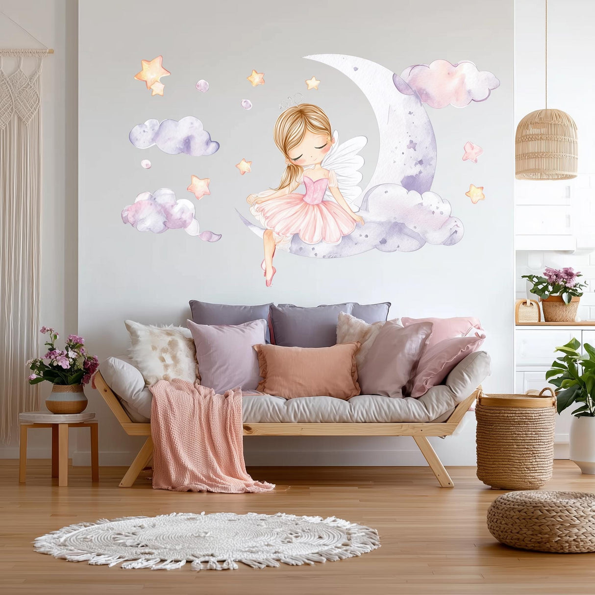 A whimsical wall decal showing a blonde fairy ballerina in a pink dress sitting on a crescent moon, surrounded by watercolor clouds and glowing stars, above a cozy white and pink-themed sofa in a nursery room.