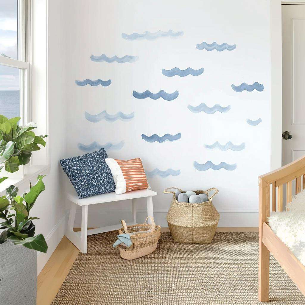 A set of blue watercolor wave decals applied randomly on a white wall in a coastal-themed kids' room with a bench, pillows, natural baskets, and light wooden furniture.
