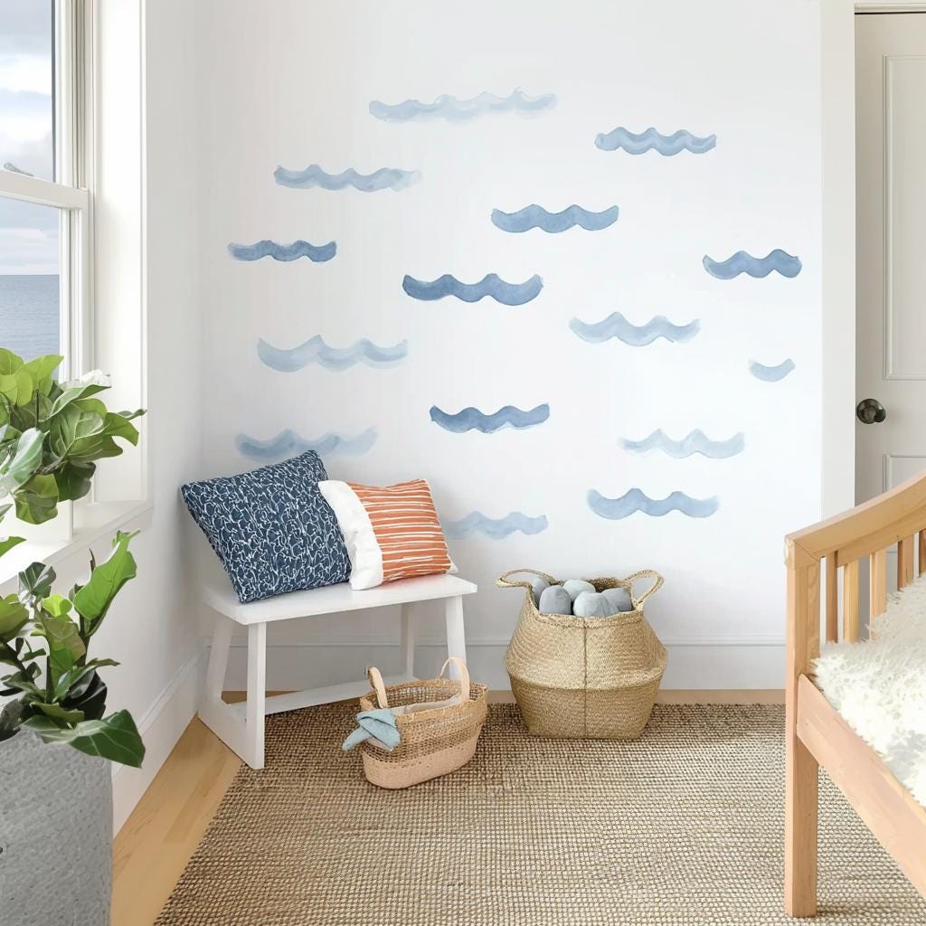 A set of blue watercolor wave decals applied randomly on a white wall in a coastal-themed kids' room with a bench, pillows, natural baskets, and light wooden furniture.