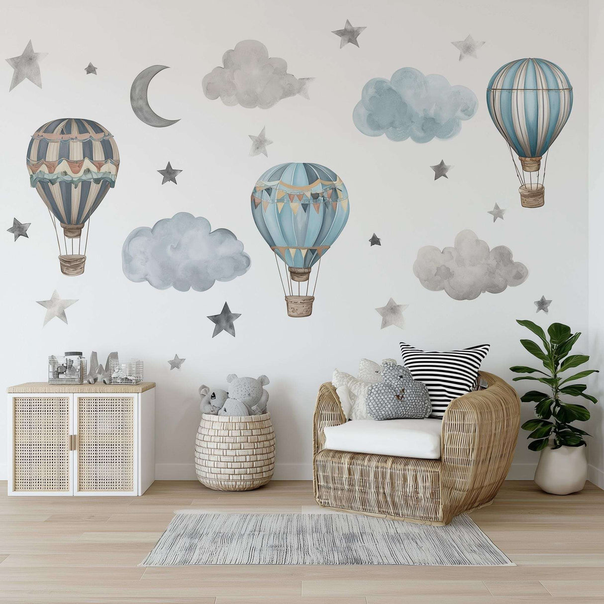 A boys’ nursery decorated with watercolor-style wall decals featuring three hot air balloons in muted blue and beige tones, surrounded by fluffy clouds, gray stars, and a crescent moon above a rattan chair and soft rug.