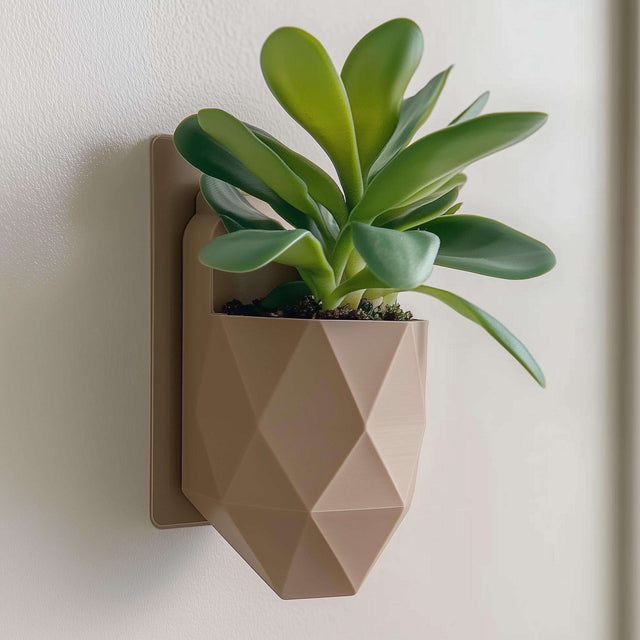 A modern wall-mounted planter with a faceted geometric design, printed in matte beige PLA and holding a small leafy green plant.