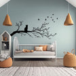 Vinyl wall decal of a cat sitting on a tree branch with flying birds, cut in solid black and applied to a pale green wall above a child's bed in a cozy, modern nursery.