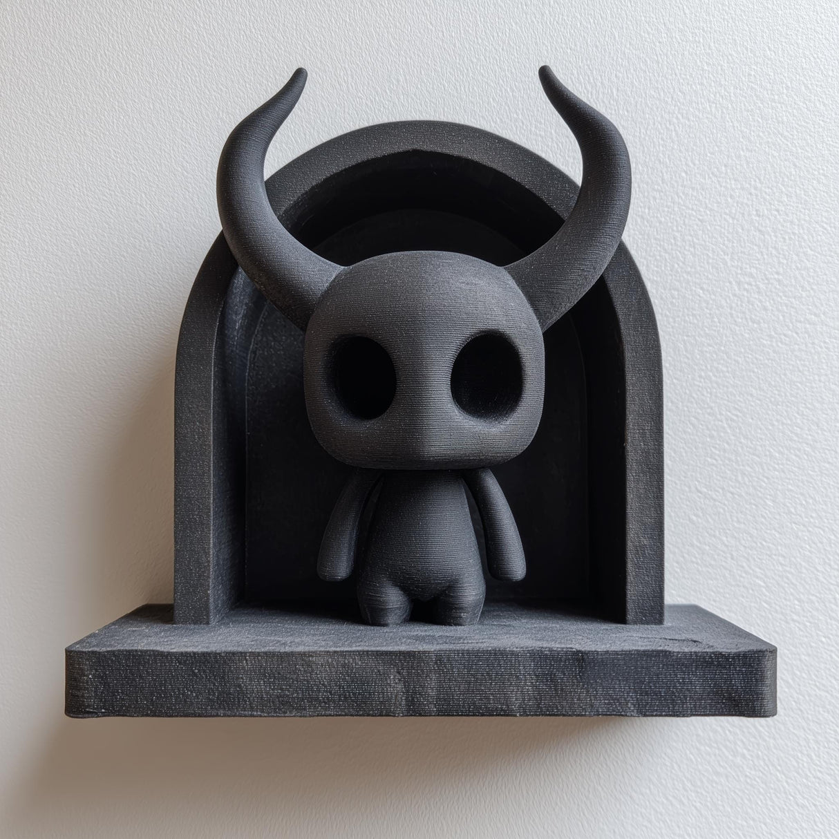 3D printed matte black wall shelf with a small horned spirit figure standing inside an arched niche, mounted on a light wall.