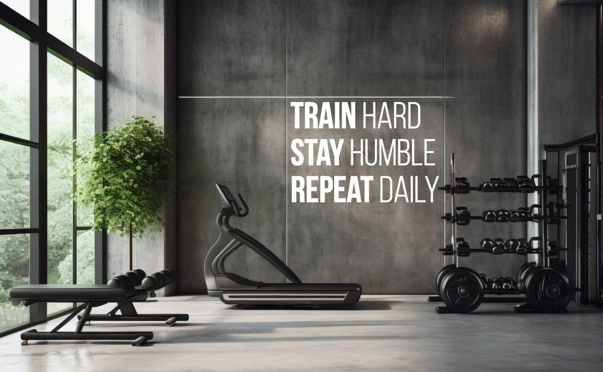 Motivational fitness wall decal in modern gym emphasizing 'Train Hard, Stay Humble, Repeat Daily' for workout inspiration.