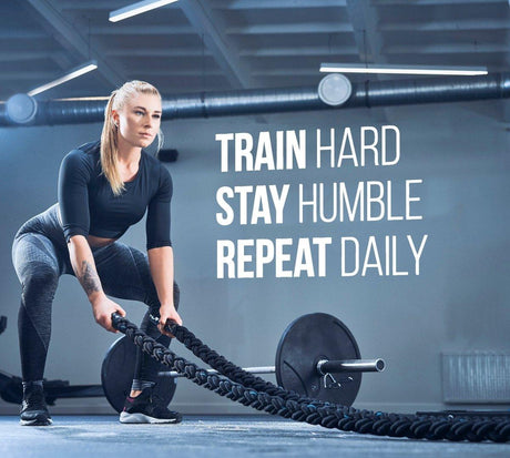 Motivational fitness wall decal sticker with the quote "Train Hard, Stay Humble, Repeat Daily" in a gym setting.