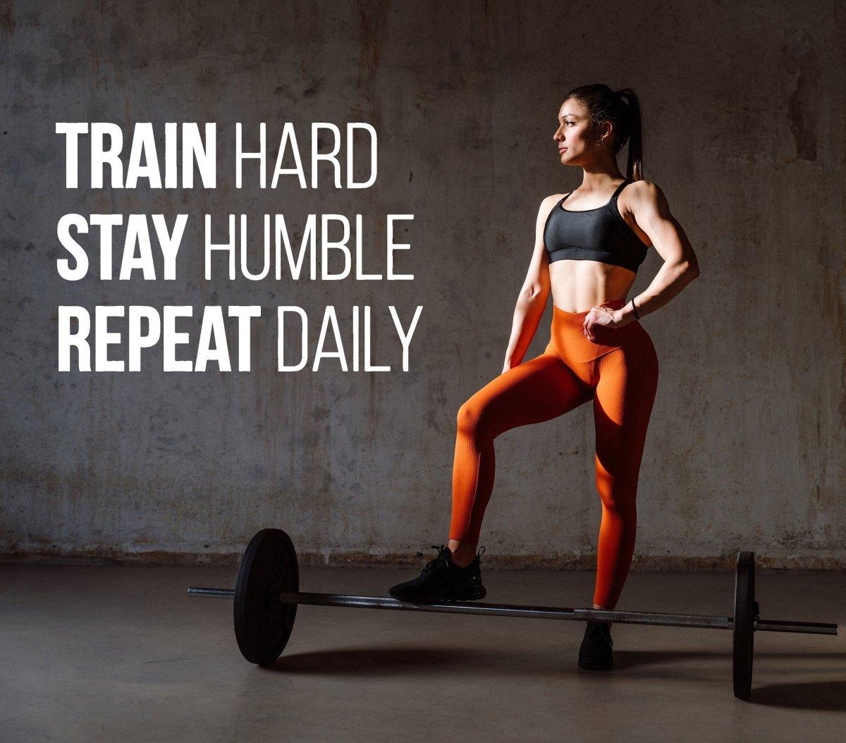 Fit woman in sporty attire beside motivational gym quote on wall decal: "Train hard, stay humble, repeat daily."