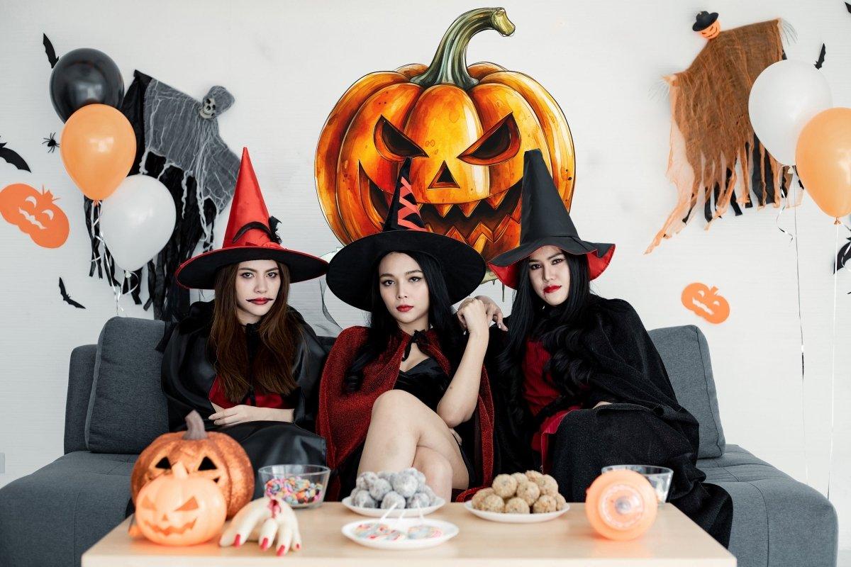 Three witches in Halloween costumes sitting on a couch, with a creepy pumpkin wall decal behind them, surrounded by holiday decor.