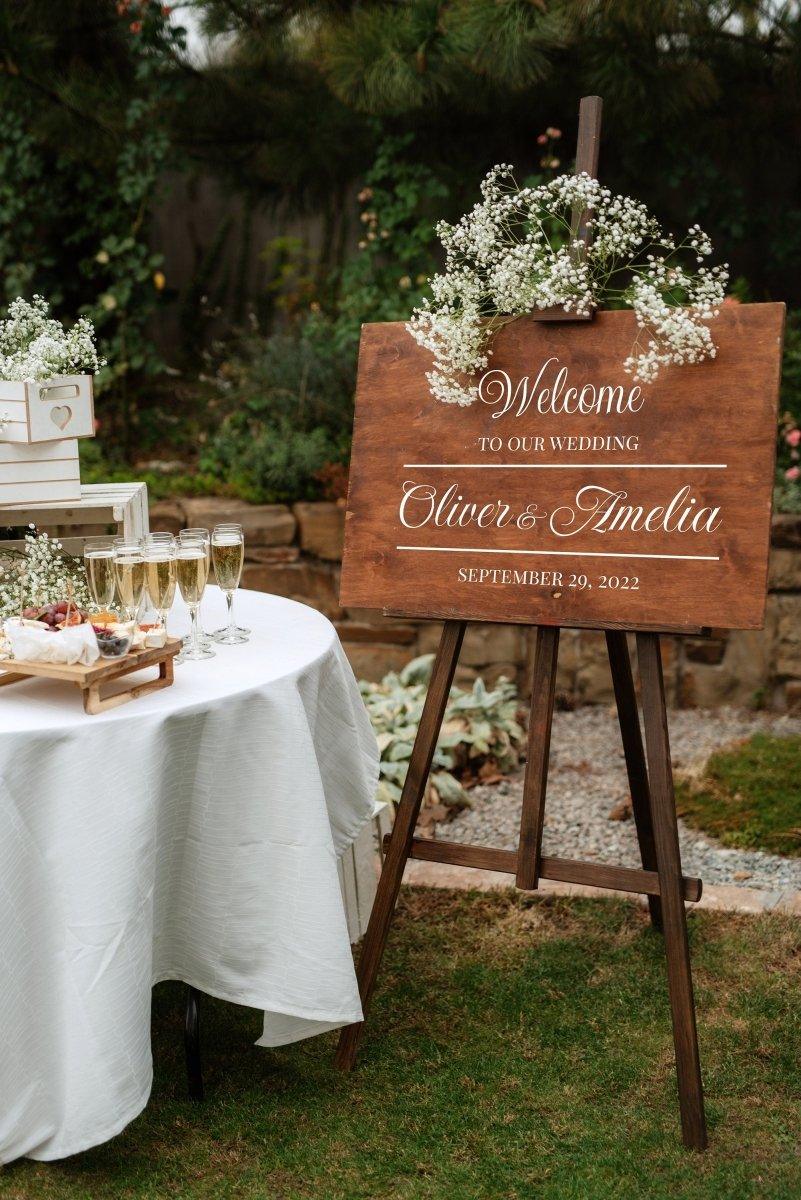 Customizable wedding welcome sign featuring couple's names and date, decorated with flowers in an outdoor setting.
