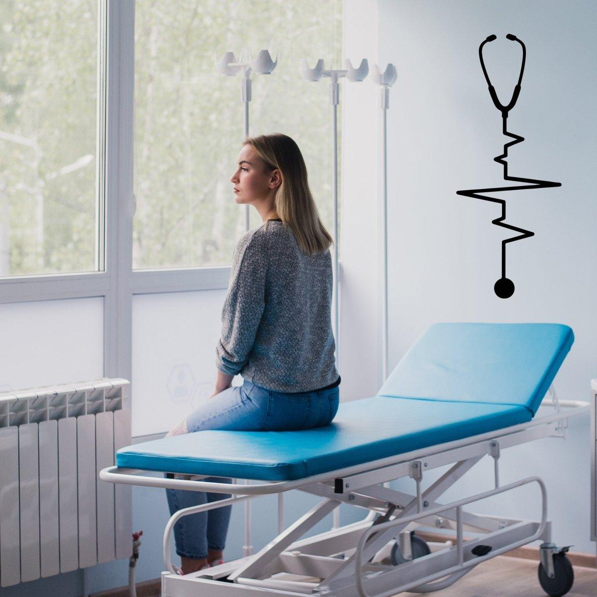 Woman sitting on a medical examination table with a stethoscope and heartbeat decal on the wall in a clinic.