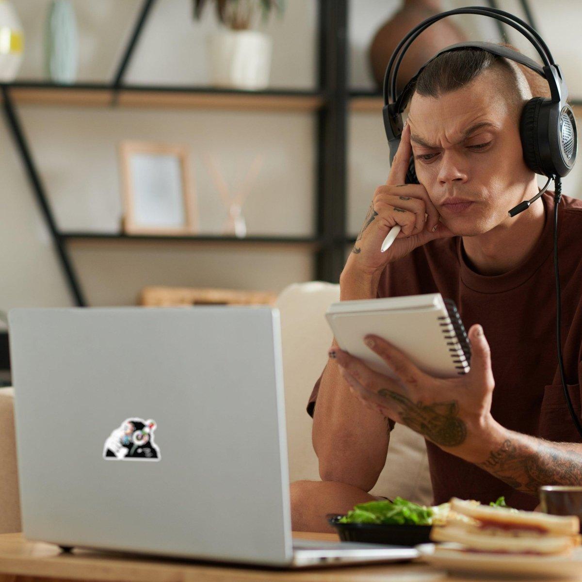 Man wearing headphones focused on laptop with luminous monkey decal sticker, taking notes while enjoying a meal.