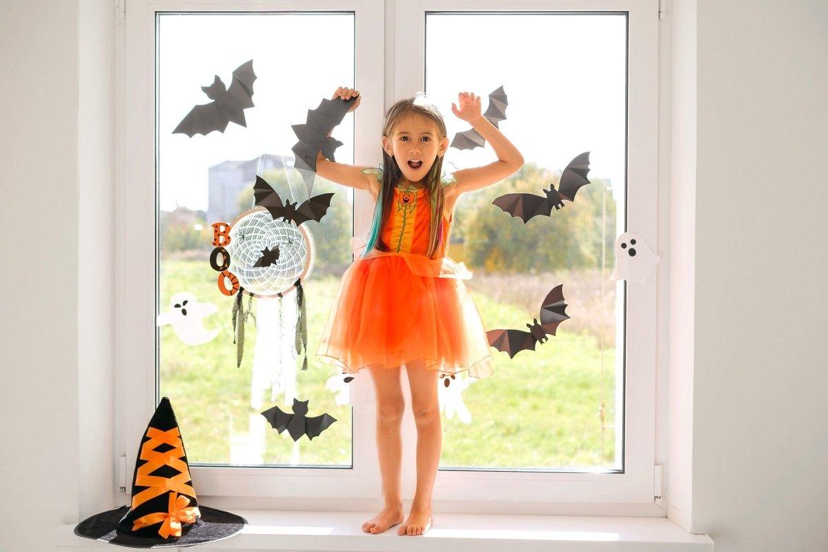 Child in orange dress playing with Halloween bat decals on window, surrounded by spooky decorations.