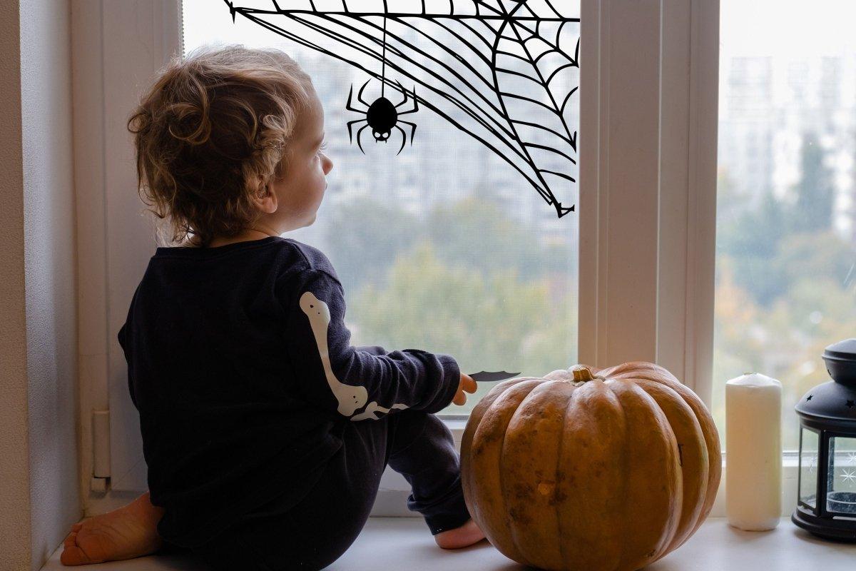 Child sitting by the window with a Halloween spider web decal and a pumpkin, creating a spooky autumn ambiance.