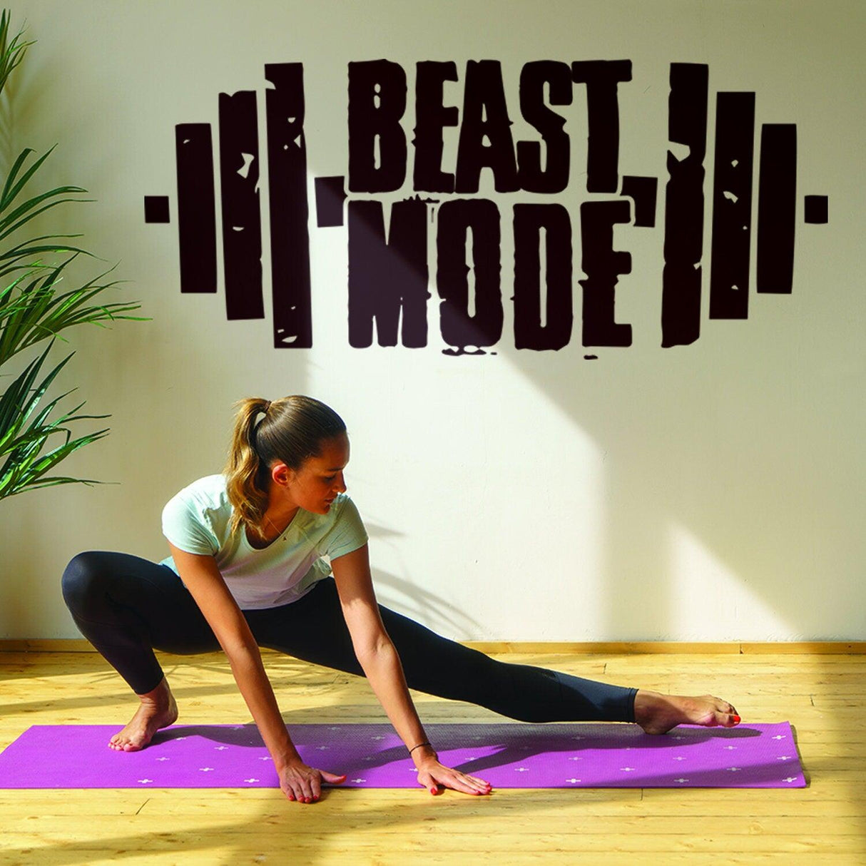 Woman stretching on yoga mat with 'BEAST MODE' vinyl decal on wall in fitness room.