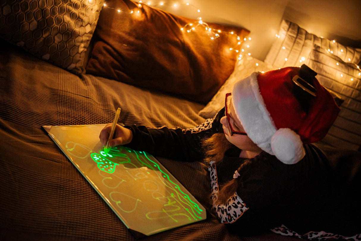 Child in Santa hat using Light Drawing Board, creating glowing neon drawings in a cozy atmosphere.