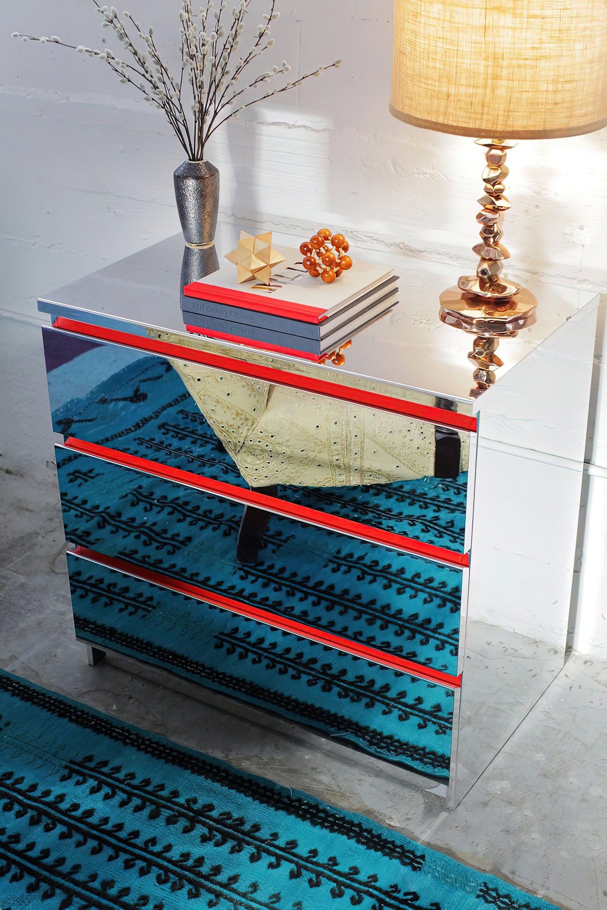 Stylish mirrored nightstand with orange accents on a blue patterned rug, featuring a lamp and decorative items.