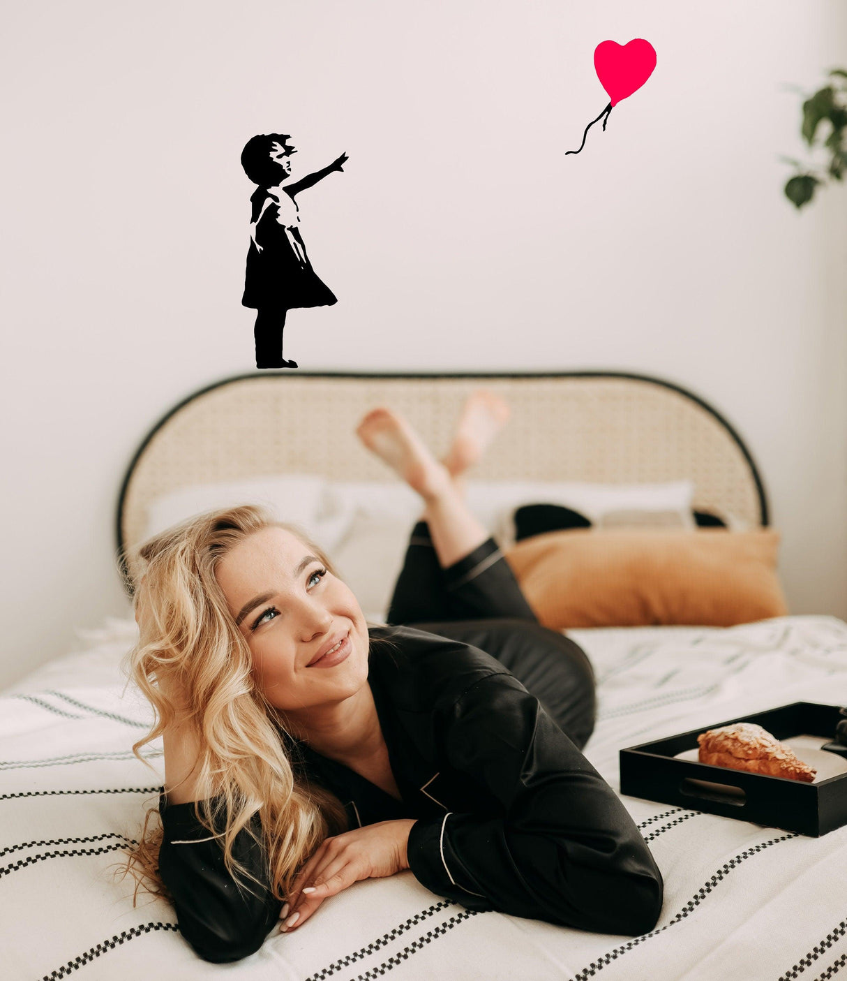 Woman relaxing on bed with Banksy girl reaching for red balloon wall decal above.