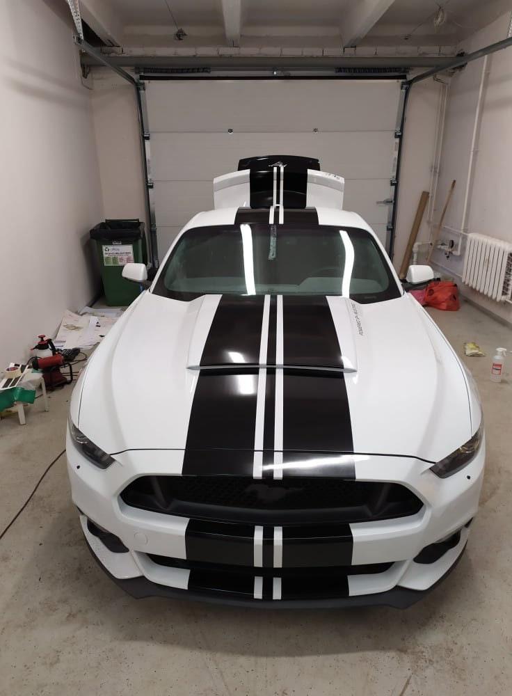 Front view of a white car with black racing stripes parked in a garage, showcasing its sporty design.