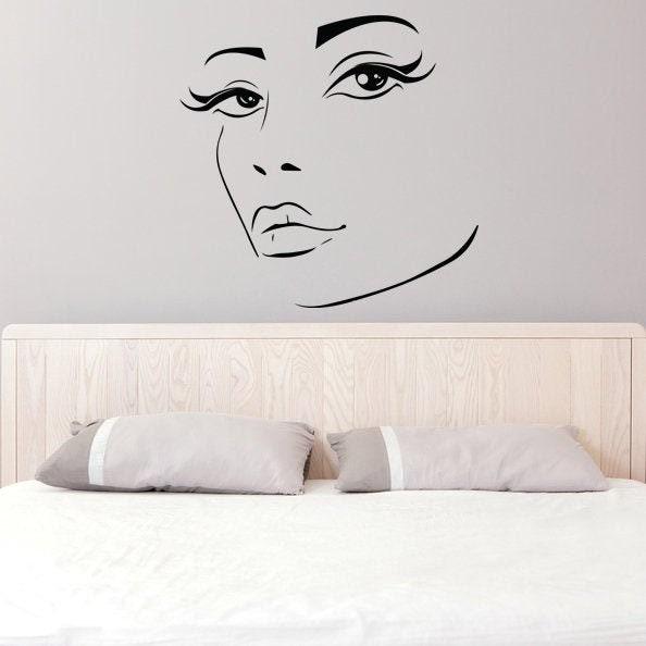 Wall decal of a stylish woman's face with long lashes above a bed, perfect for feminine bedroom decor.