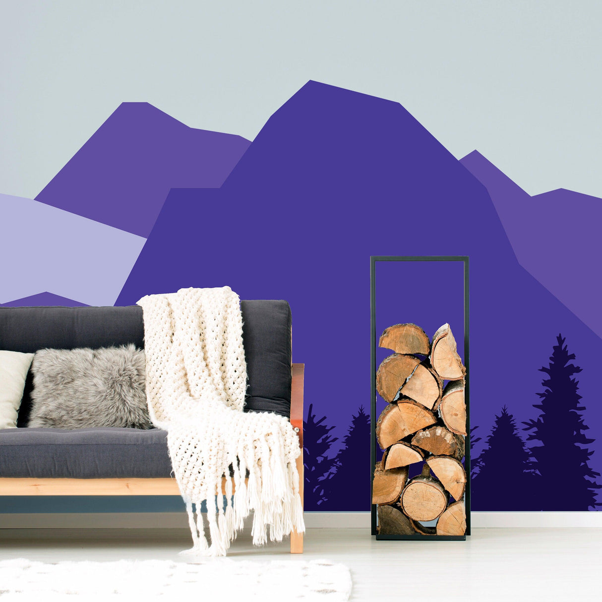 Mountain wall decal in purple hues above a cozy sofa with a knitted throw and a log holder.