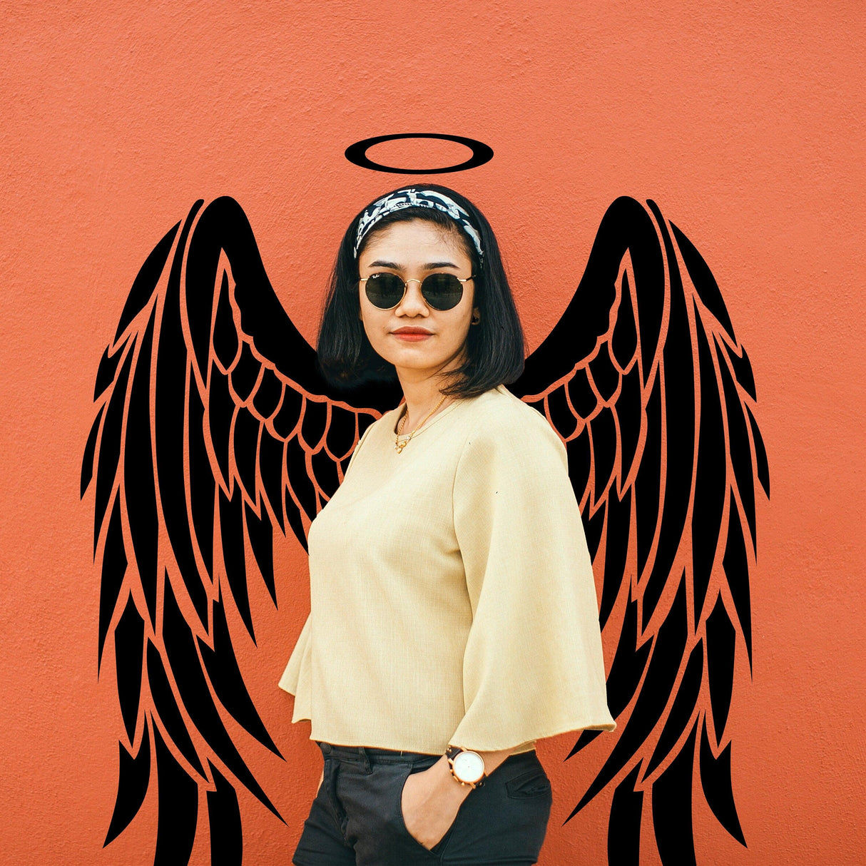 Woman posing in sunglasses with angel wings decal on an orange wall, embodying elegance and whimsy.