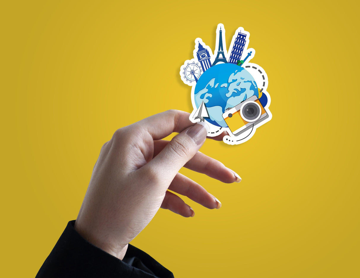 Hand holding a colorful travel-themed vinyl sticker with a globe, landmarks, and a camera on a yellow background.