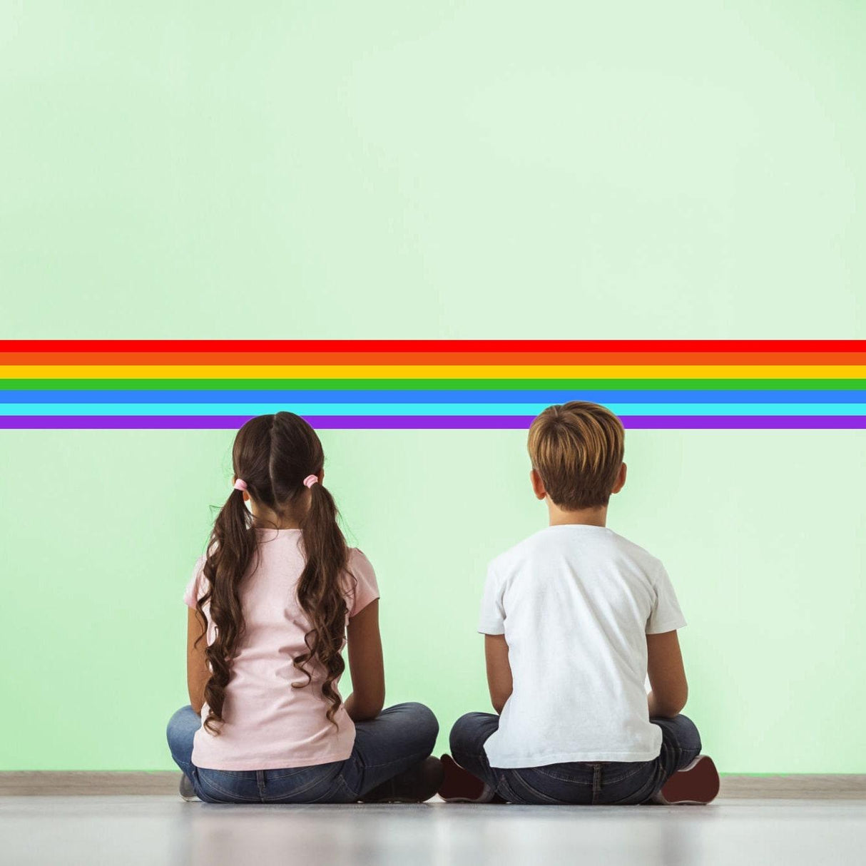 Two children sitting on the floor, admiring colorful rainbow border sticker on a green wall.