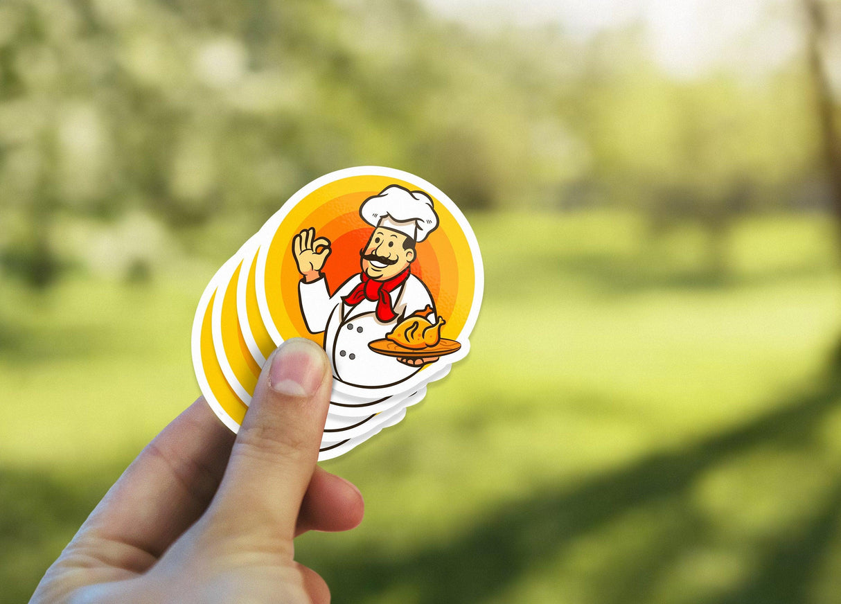 Hand holding colorful chef stickers outdoors, featuring a friendly chef with a baked dish design.