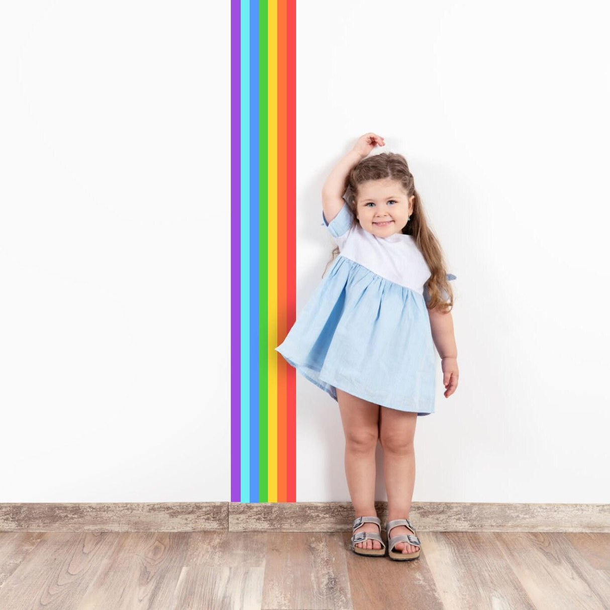 Smiling girl in blue dress standing next to colorful rainbow stripe wall decal, perfect for nursery decor.