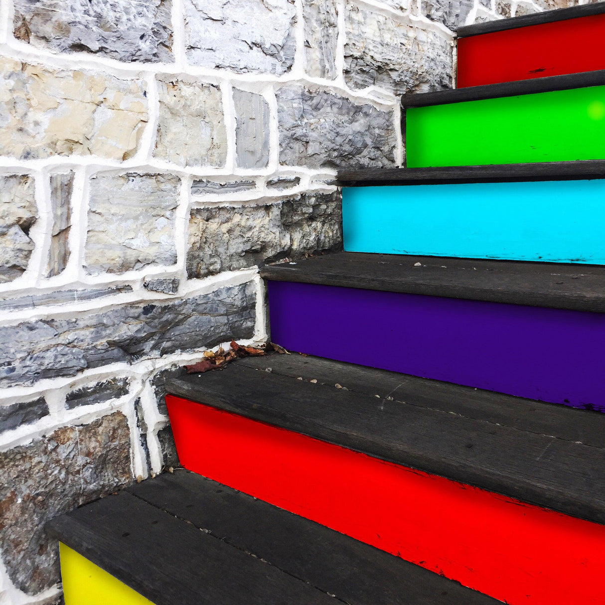 Colorful rainbow stair riser stickers applied on wooden steps, adding vibrant décor to a stone wall staircase.