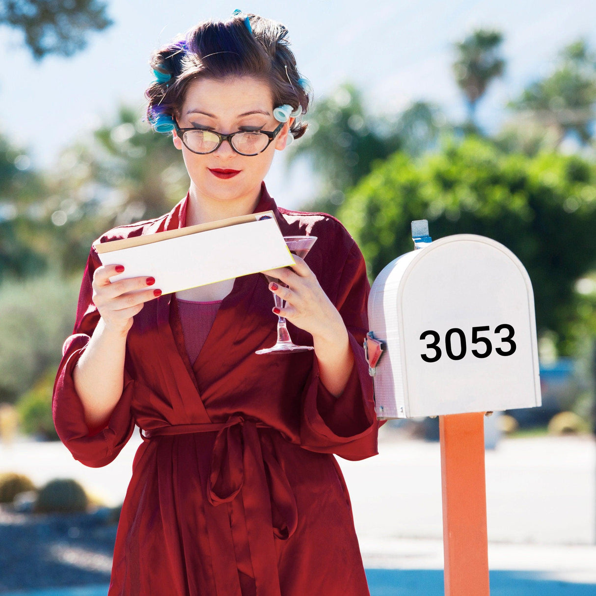 Person in a red robe checking mailbox with custom address decal, styled with vintage curls and sipping a drink.