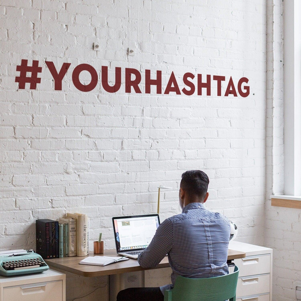 Person working at a desk with a personalized hashtag vinyl sticker on the wall, showcasing #YOURHASHTAG.