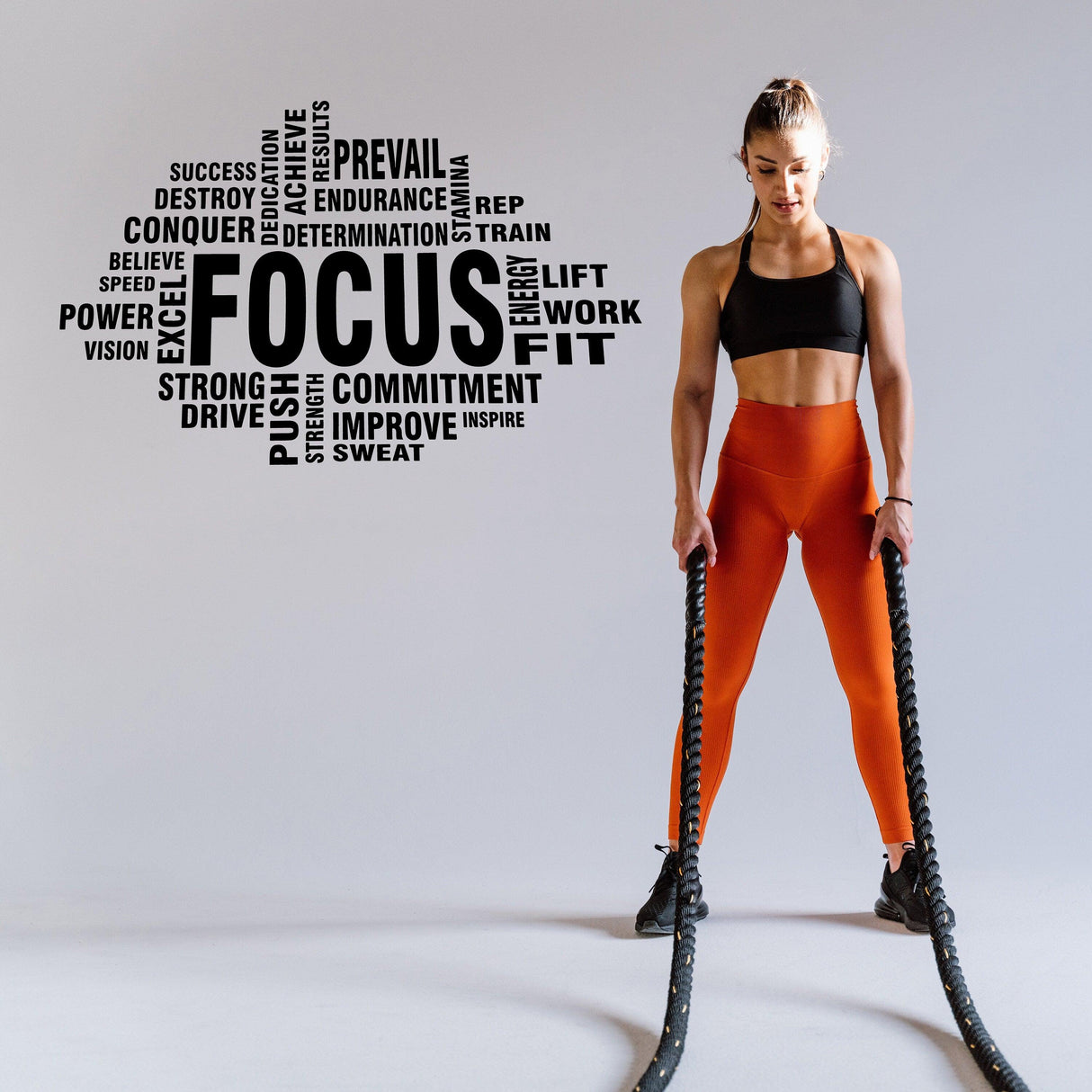 Fitness enthusiast lifting battle ropes with motivational words on wall, showcasing strength and focus during workout.