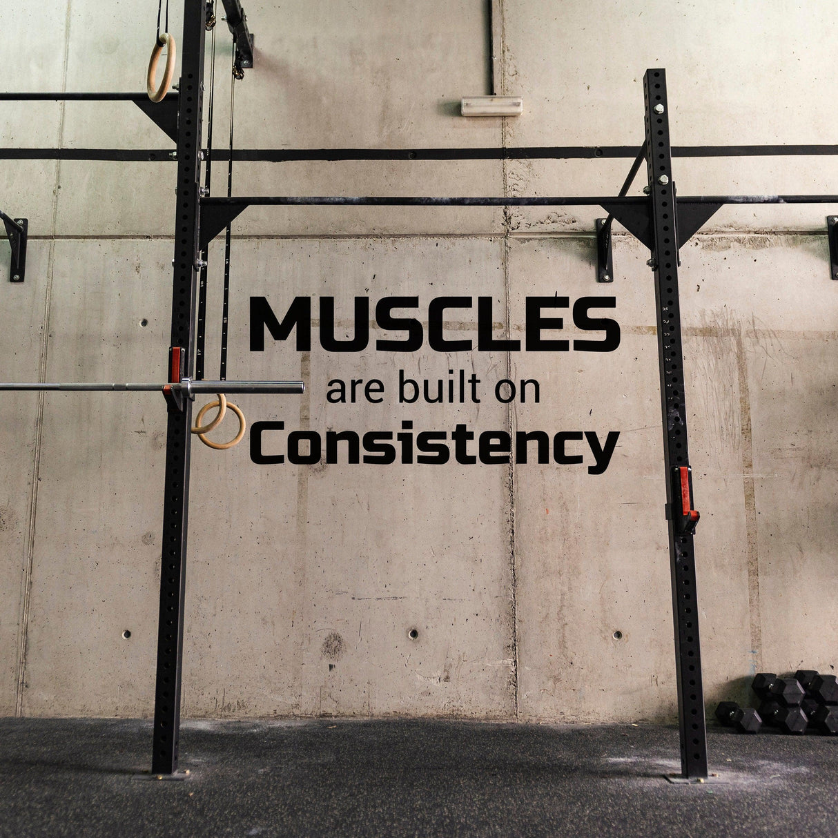 Fitness inspiration wall sticker with quote 'Muscles are built on consistency' in a gym setting.