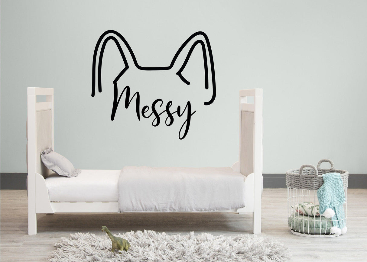 Vinyl wall decal of cat ears with the word 'Messy' above a child's bed in a cozy room.