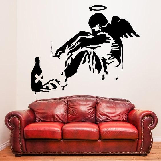 Banksy Fallen Angel wall sticker above a red leather sofa in a modern living room setting.