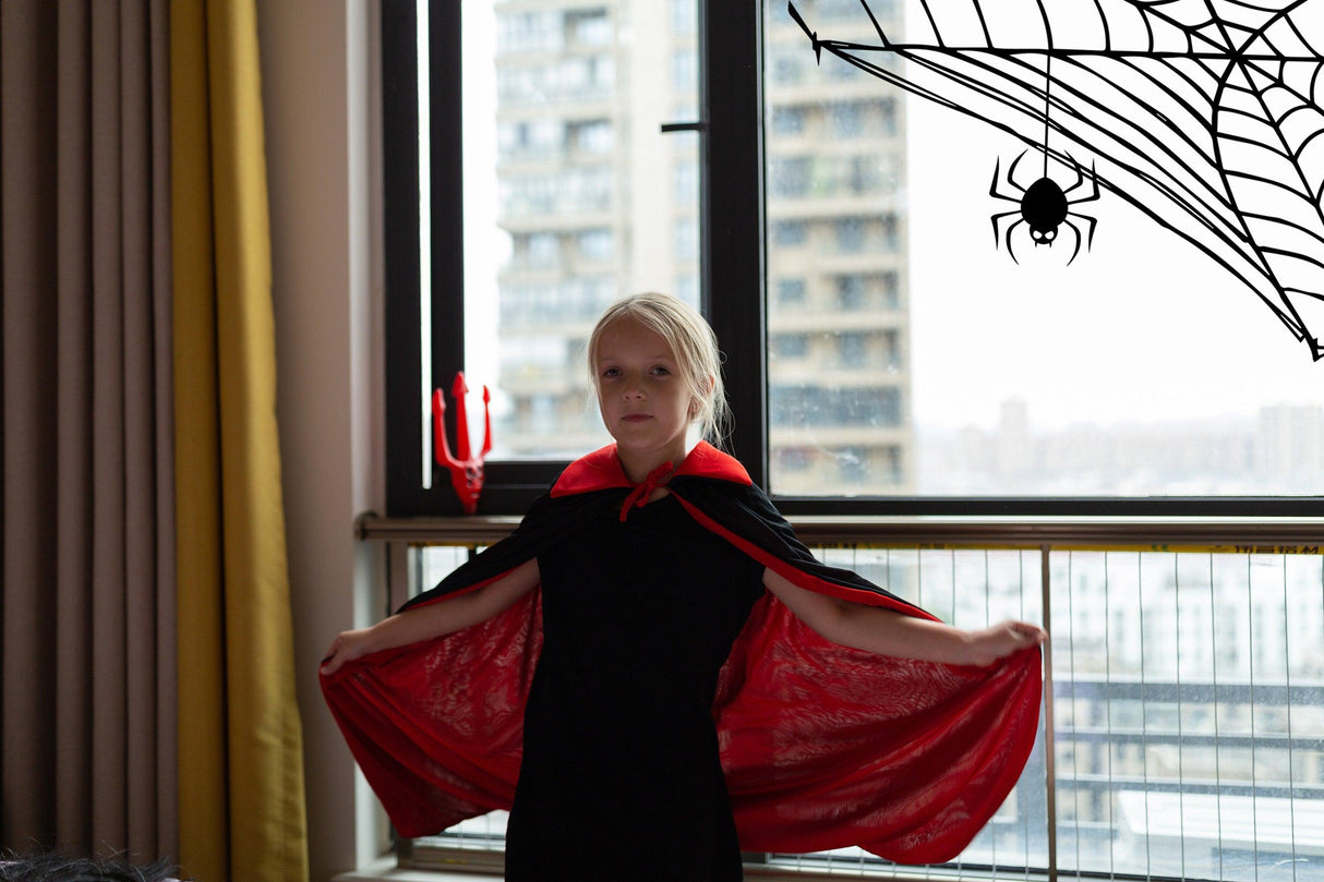Child wearing a black outfit and red cape with Halloween spider web decals on window behind them.