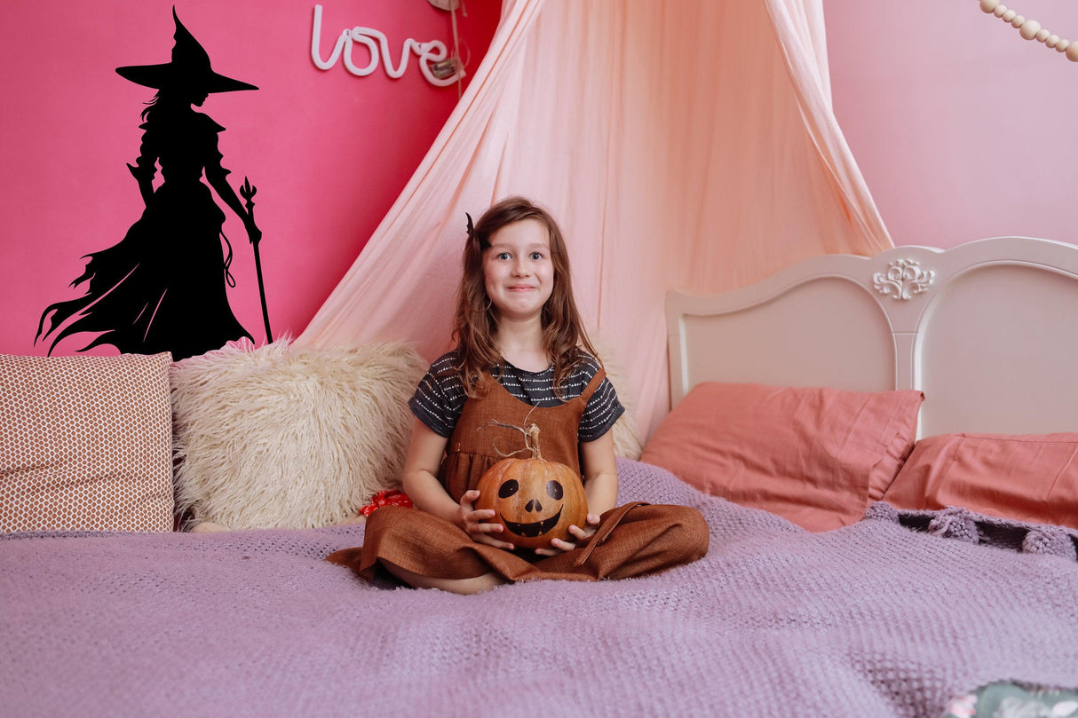 Girl sitting on a bed holding a pumpkin with a Wicked Witch Silhouette decal in the background for Halloween decor.