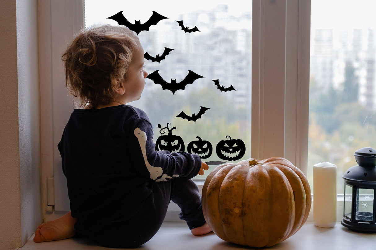 Child sitting by the window decorated with Halloween bats and pumpkins decals, alongside a pumpkin and candle.