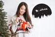 Woman in a cozy sweater holding a gift with a Santa and reindeer wall decal in the background.