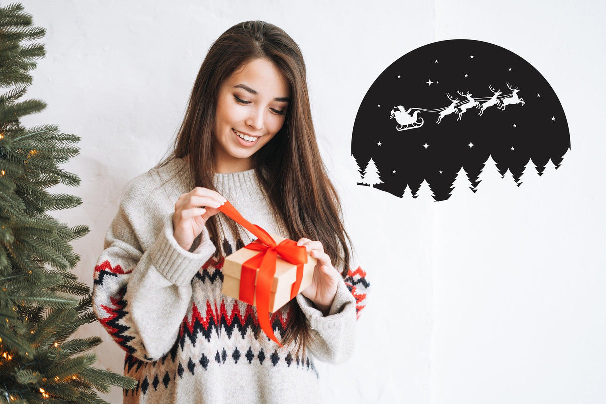 Woman in a cozy sweater holding a gift with a Santa and reindeer wall decal in the background.