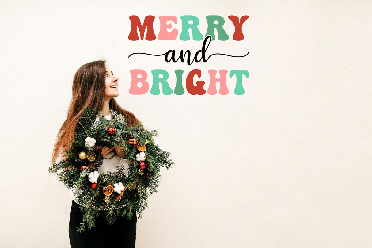 Cheerful woman holding a Christmas wreath under the text 'Merry and Bright' wall decal in festive colors.